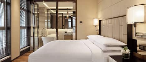 A modern hotel room with a large bed, a bedside table with a lamp, a small table with a vase of flowers, and a view of the bathroom through a glass door.
