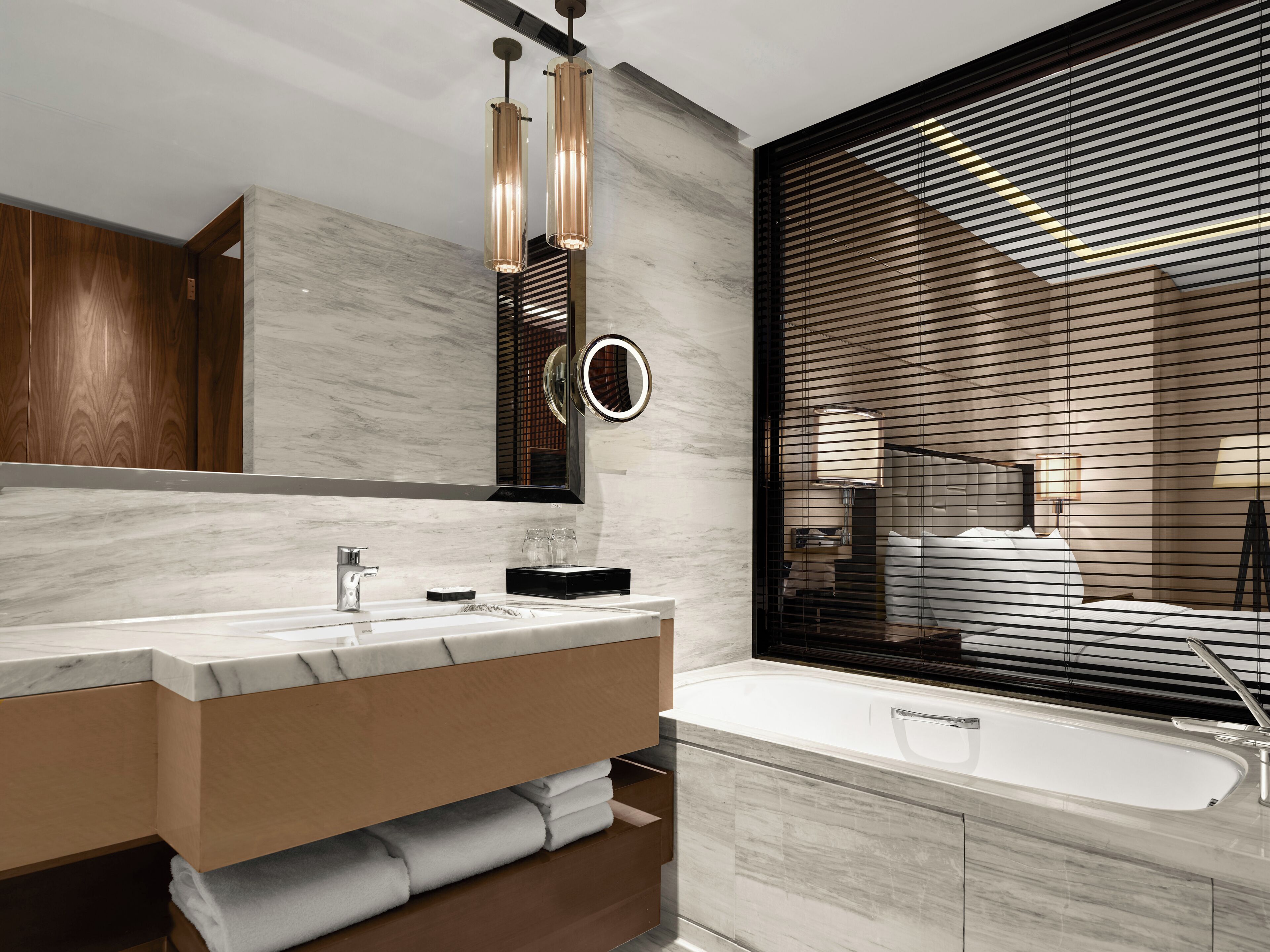 A modern bathroom with a marble vanity, a large mirror, a round mirror, a bathtub, and a window with horizontal blinds.