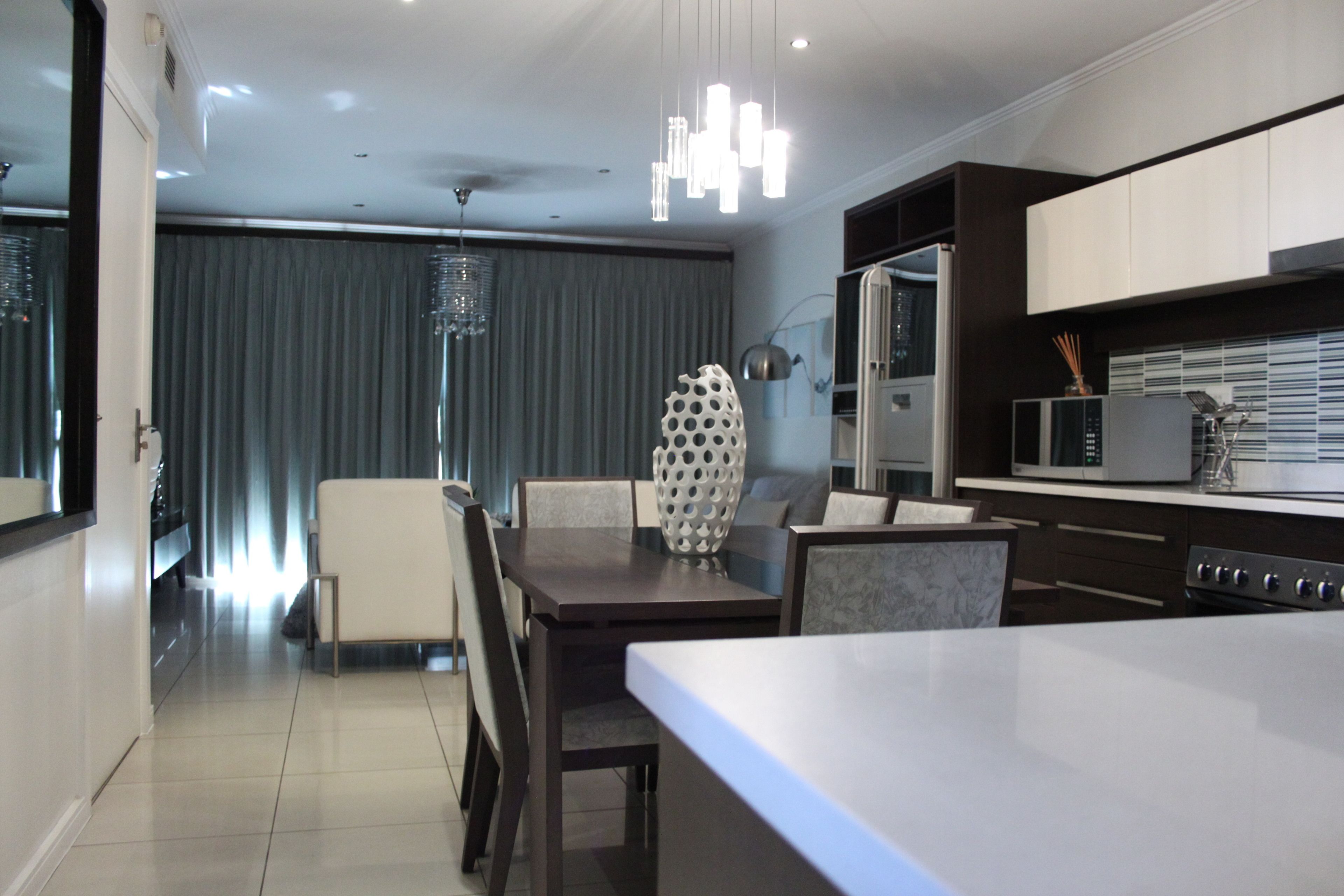 A modern kitchen with a dining area, featuring a large island, dark cabinetry, and contemporary lighting fixtures.