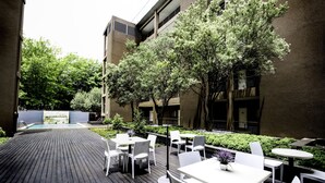 An outdoor patio area with wooden decking, white tables and chairs, and a pool in the background. - The Nicol Hotel and Apartments (Germiston)