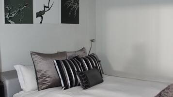 Una cama bien hecha con ropa blanca y varias almohadas, un sofá gris, y tres obras de arte colgadas en la pared que representan ramas.