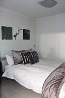 Una cama bien hecha con ropa blanca y varias almohadas, un sofá gris, y tres obras de arte colgadas en la pared que representan ramas.