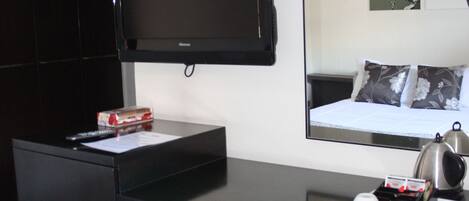 Habitación de hotel con escritorio negro, una silla, televisor de pantalla plana montado en la pared, cama con colcha floral blanco y gris, y una mesita con hervidor y tazas.