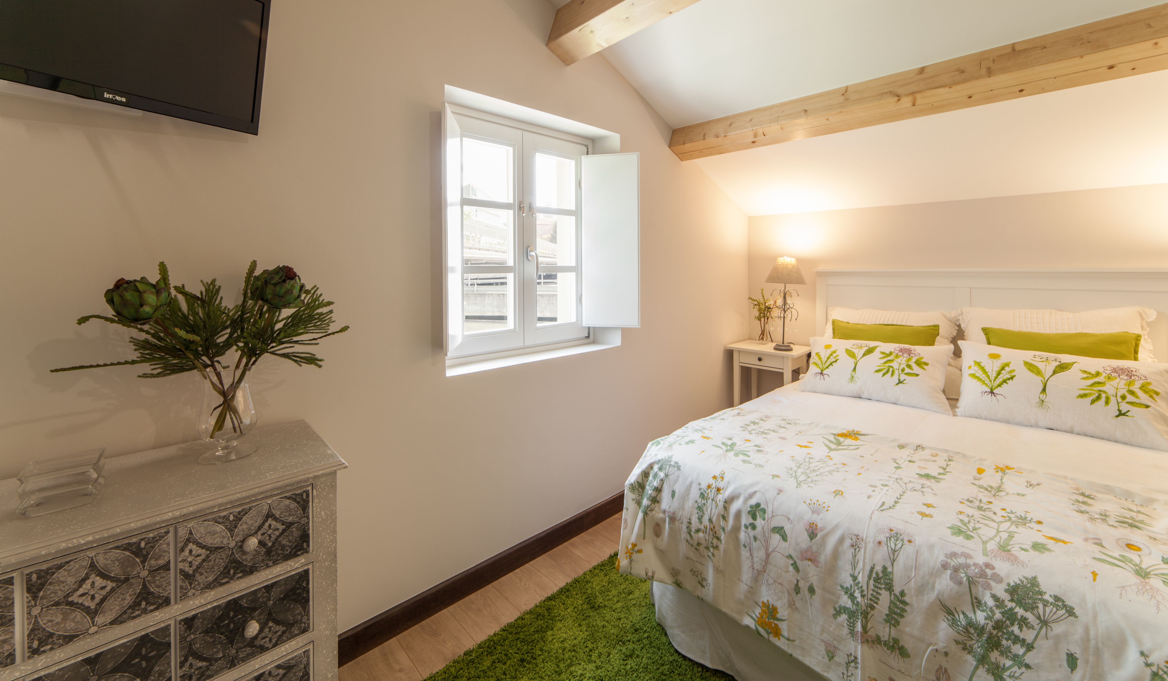Een slaapkamer met een bed, nachtkastje, lamp, televisie en een raam met uitzicht op gebouwen.