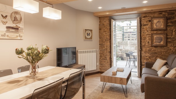 A modern living room with a dining area, stone wall, wooden beams, and a television. - Apartamentos Nova Compostela (Santiago de Compostela)