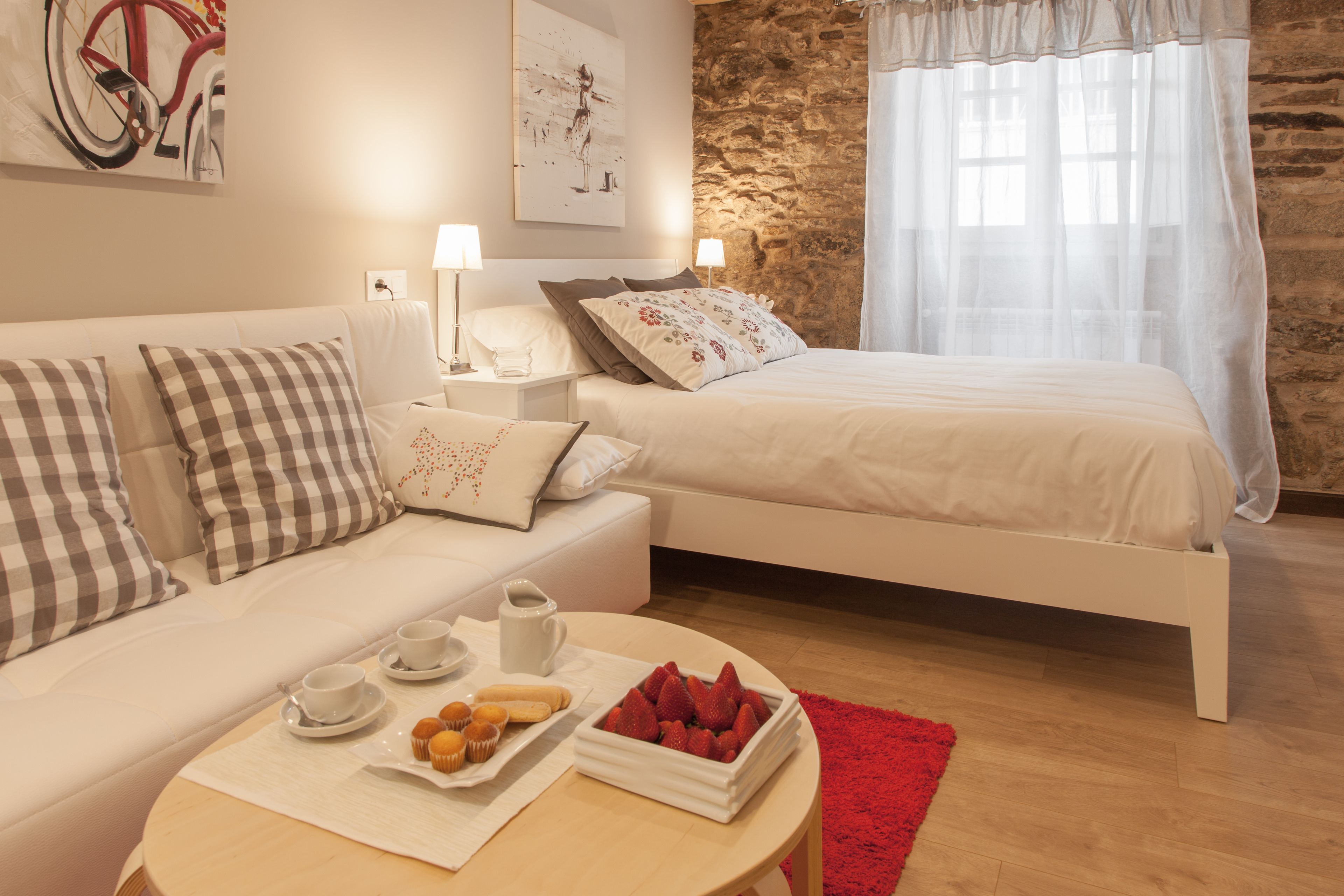 Ein modernes Schlafzimmer mit einem weißen Bett, einem weißen Sofa und einem roten Teppich. Auf einem runden Tisch steht ein Frühstücksbrett mit Muffins und Erdbeeren.