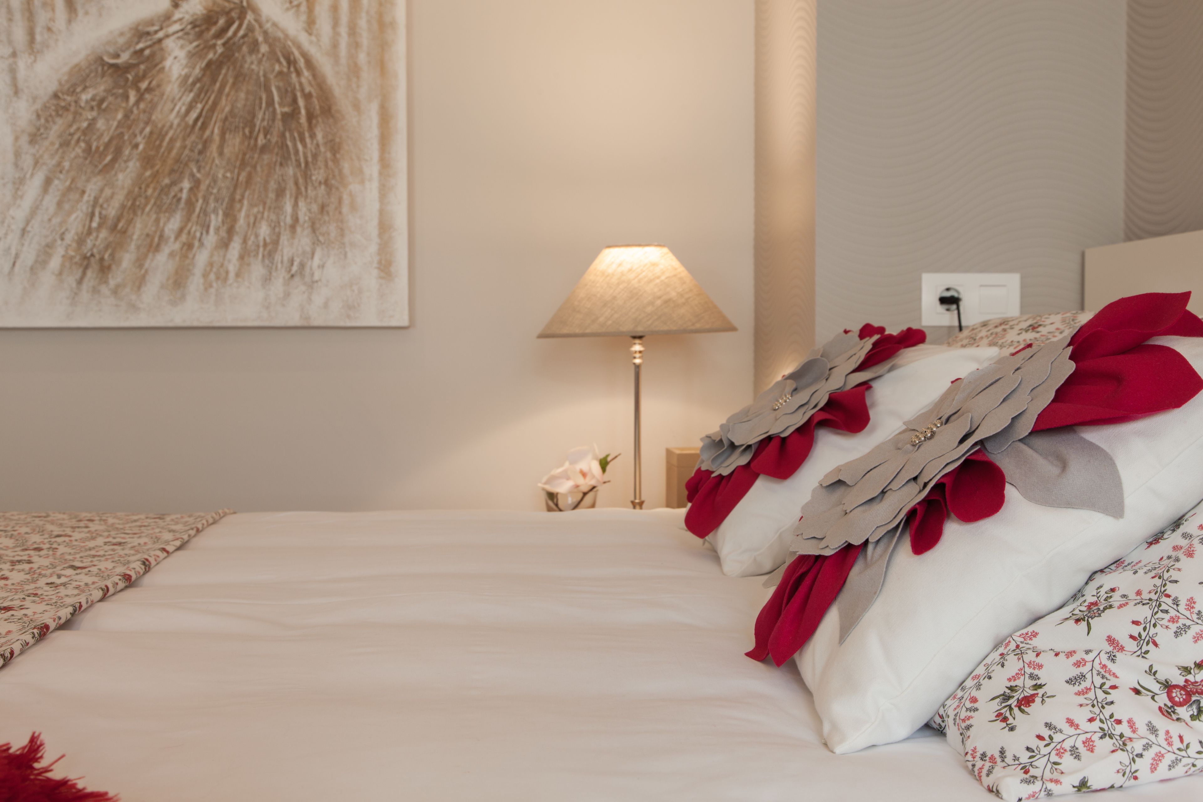 A neatly made bed with white linens, a red and white floral pillow, and a lamp on the side.
