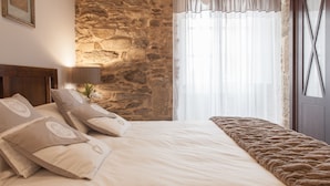 A bedroom with a stone wall, a bed with beige bedding and pillows, a bedside lamp, and a window with sheer curtains. - Apartamentos Nova Compostela (Santiago de Compostela)