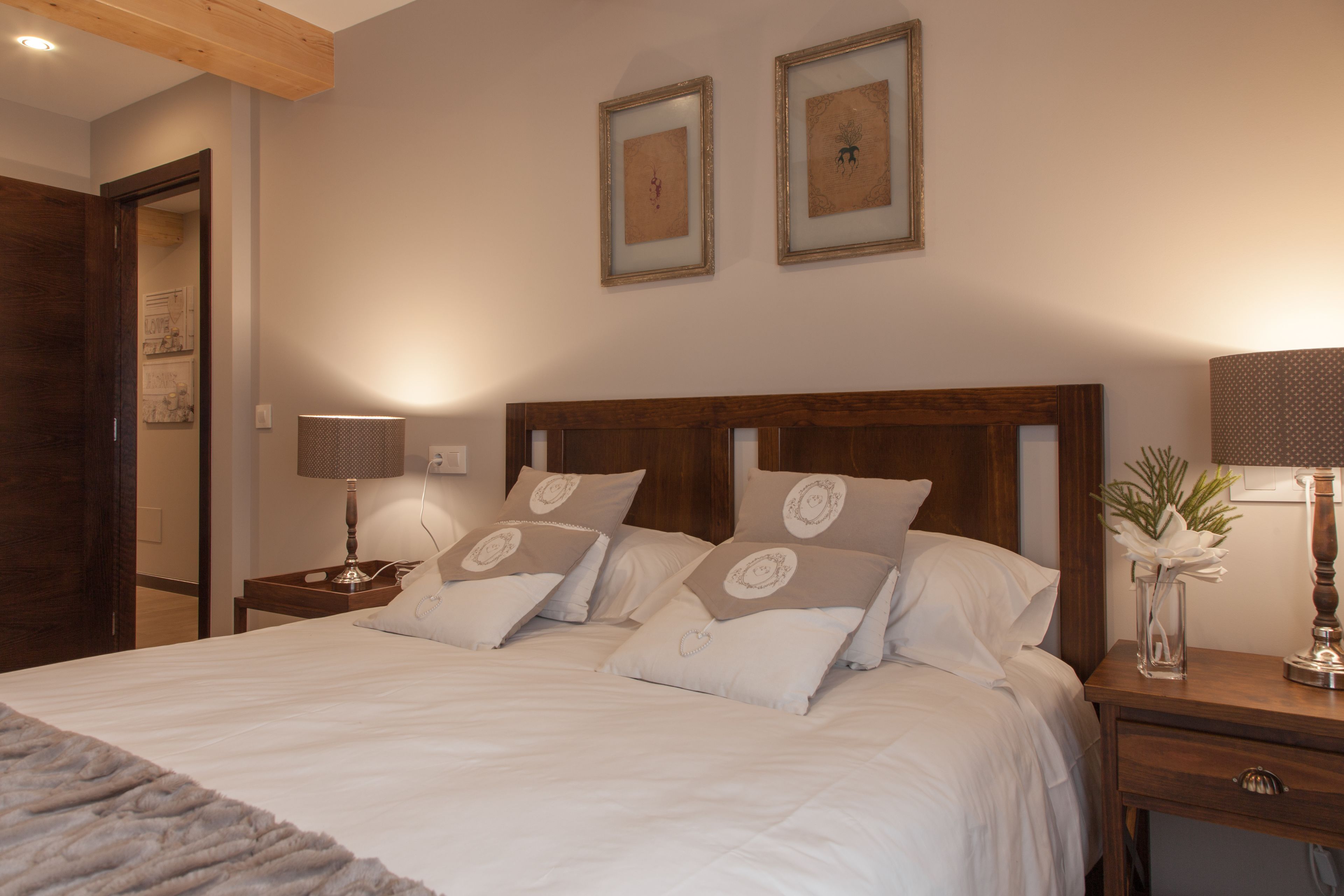 A neatly made bed with white linens and decorative pillows, a wooden headboard, a bedside table with a lamp, and framed artwork on the wall.