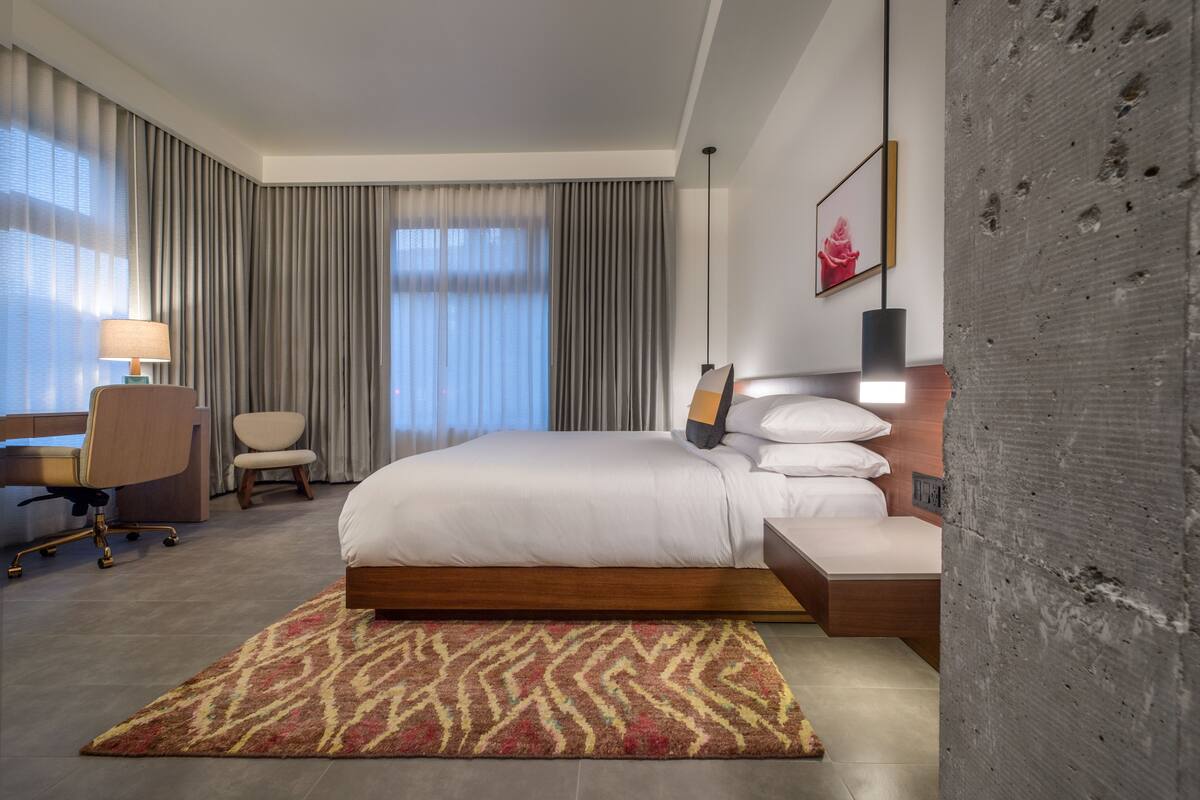 A modern hotel room with a large bed, a desk with a chair, a desk lamp, a patterned rug, and a large window with curtains.