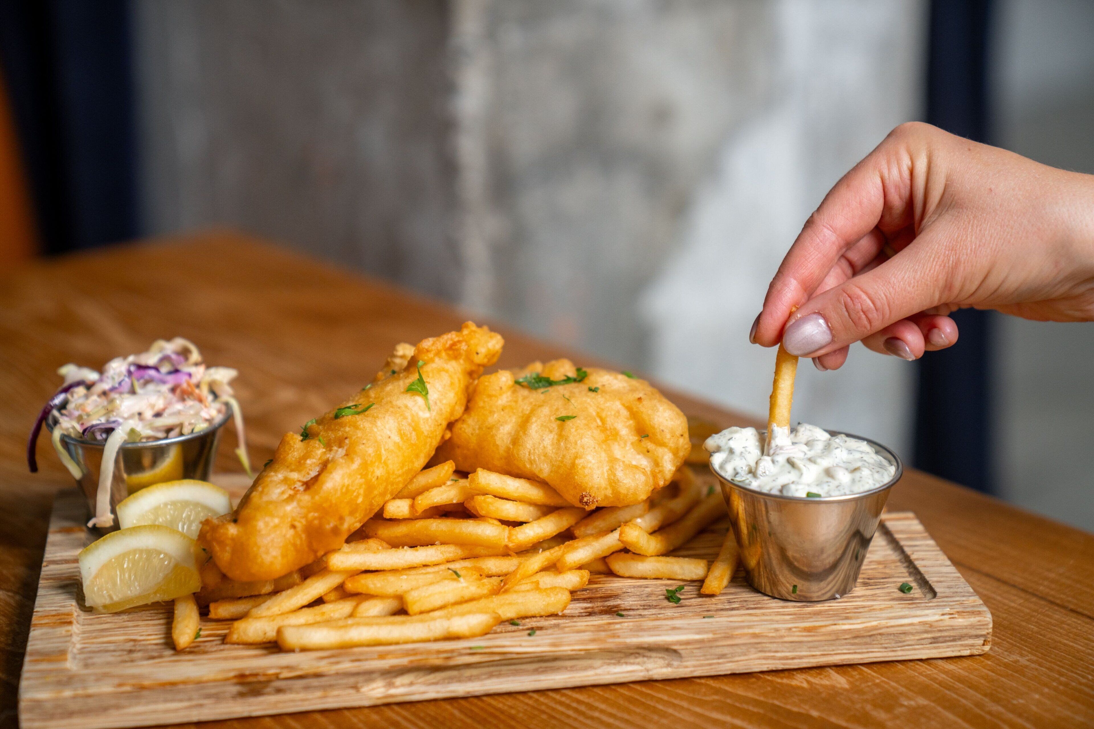 A wooden table with a meal consisting of fish and chips, a small metal cup of tartar sauce, and a side of coleslaw.