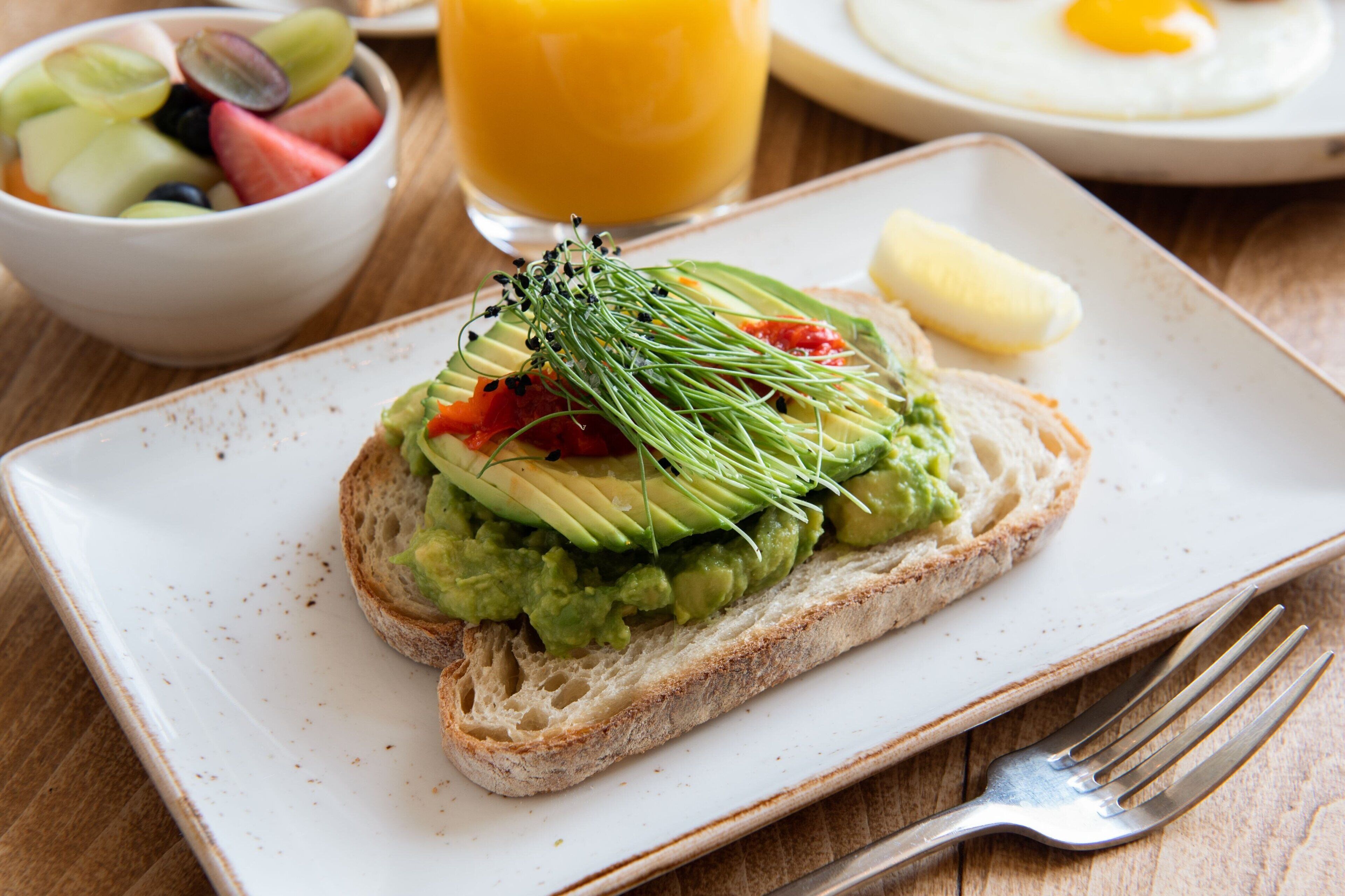 A plate with avocado toast, a bowl of fruit, a glass of orange juice, and a plate with a fried egg.