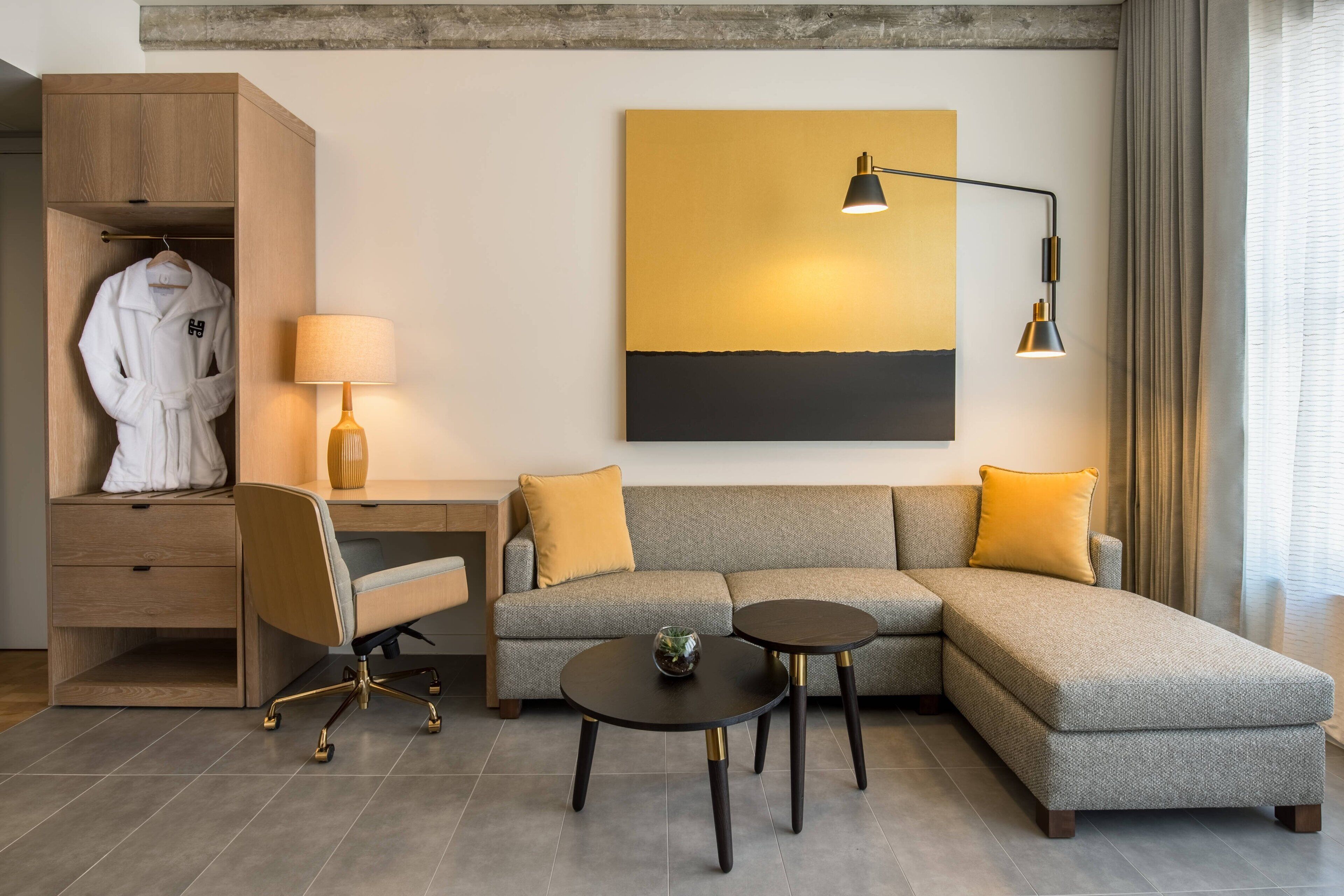 Une chambre d’hôtel moderne avec un canapé d’angle gris, une armoire en bois, un bureau avec une lampe et une table basse ronde.