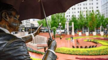 A statue of a man holding an umbrella in an urban setting with a circular garden and buildings in the background.