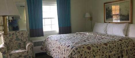 A hotel room with a large bed, a chair, a window with curtains, a painting on the wall, and a lamp.