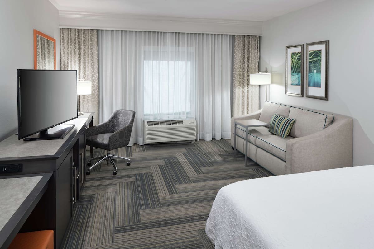 Une chambre d’hôtel comprenant un lit, un bureau avec une télévision, une chaise, un canapé et une fenêtre avec des rideaux.