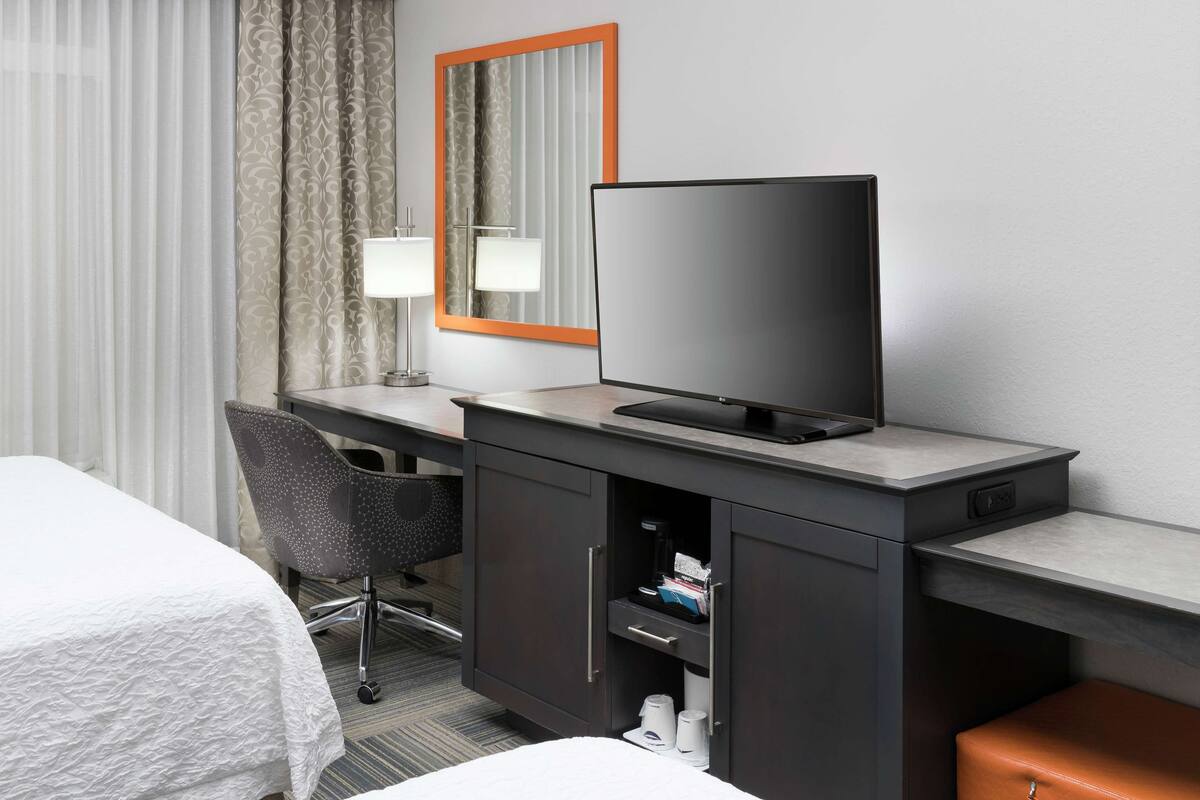 Une chambre d’hôtel comprenant un lit, un bureau avec une télévision, une chaise, un meuble de rangement et une fenêtre avec des rideaux.