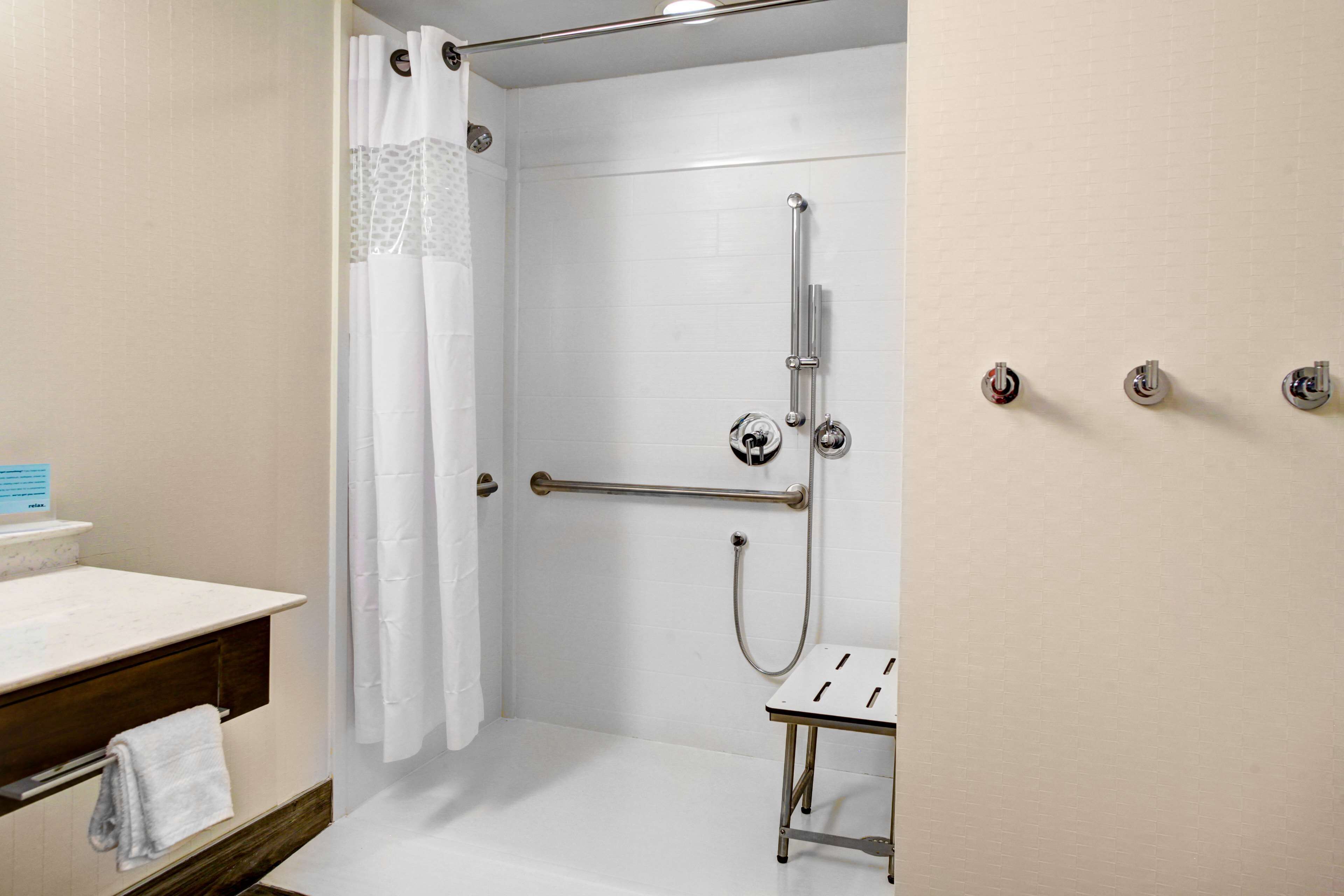 A shower area with a white tiled wall, a showerhead, a handrail, and a small table.