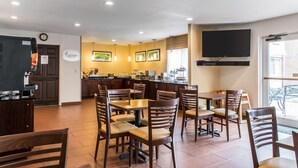 Un restaurant avec des tables et des chaises en bois, un espace cuisine et un téléviseur à écran plat fixé au mur.