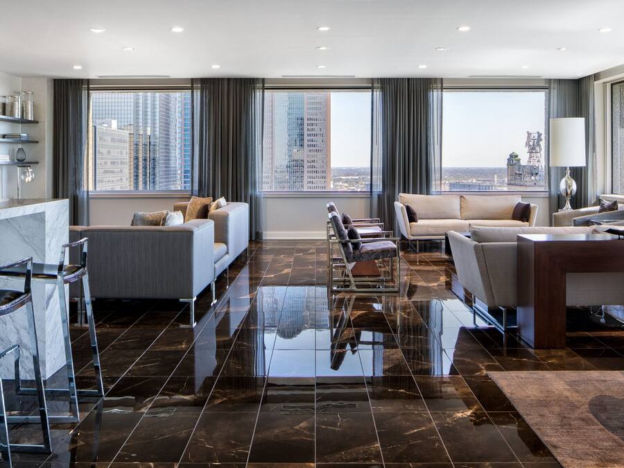 Una habitación de hotel moderna con un ventanal grande que ofrece vistas a la ciudad, una zona de estar con sofás y sillones, y un suelo de baldosas de mármol.