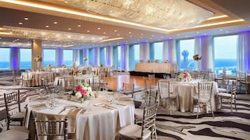 A spacious banquet hall with round tables, elegant chandeliers, and a view of the ocean.