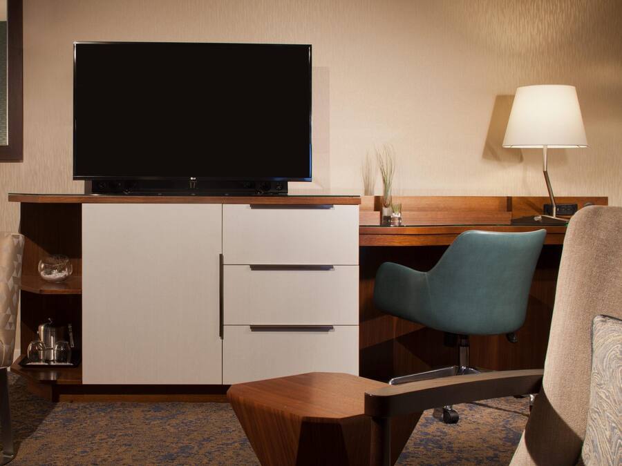 Habitación de hotel con televisión de pantalla plana montada en un mueble, un escritorio con lámpara, dos sillas y una cafetera en la mesita de noche.