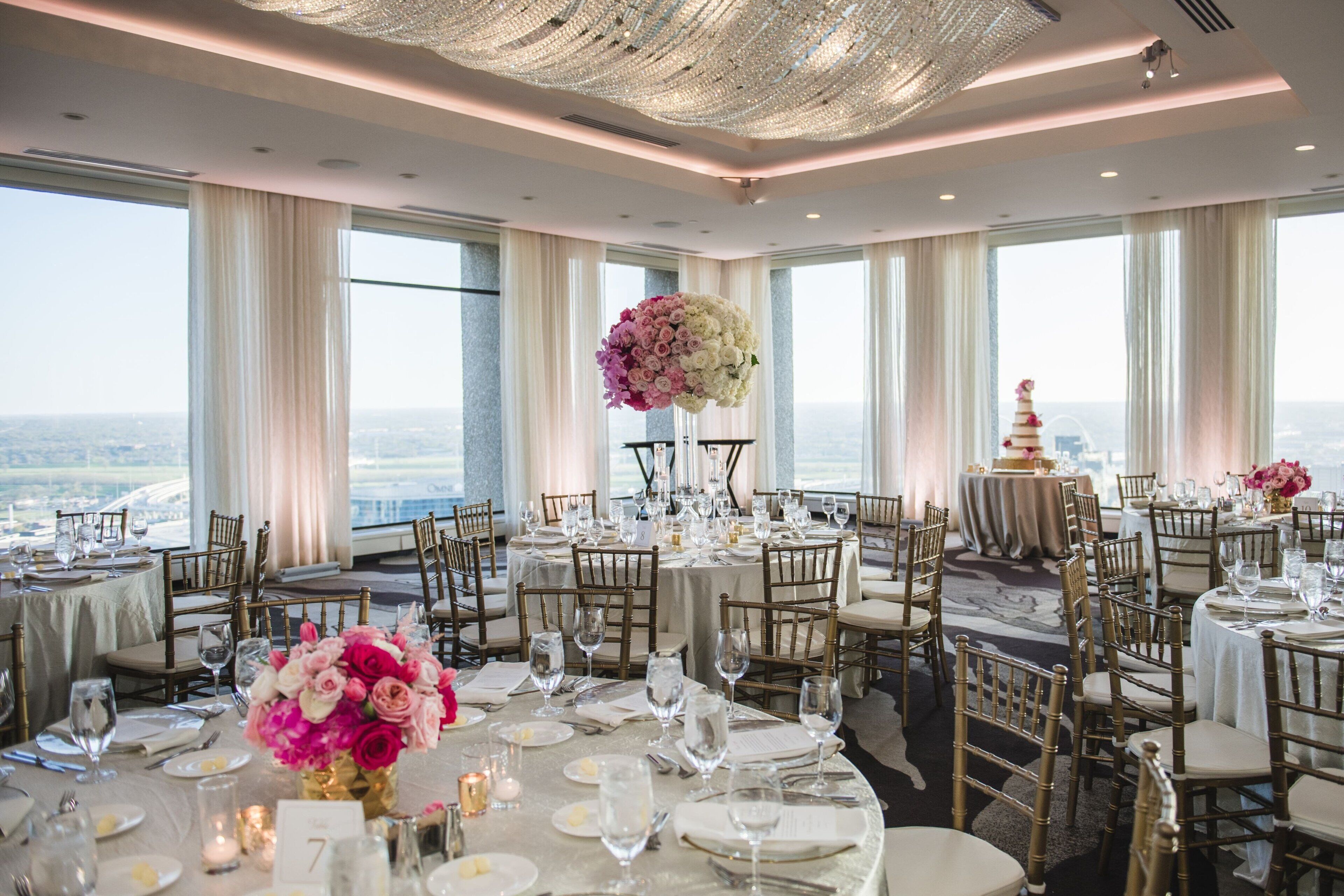 A banquet hall with round tables set for a formal event, large floral arrangements, and a view of the city through large windows.
