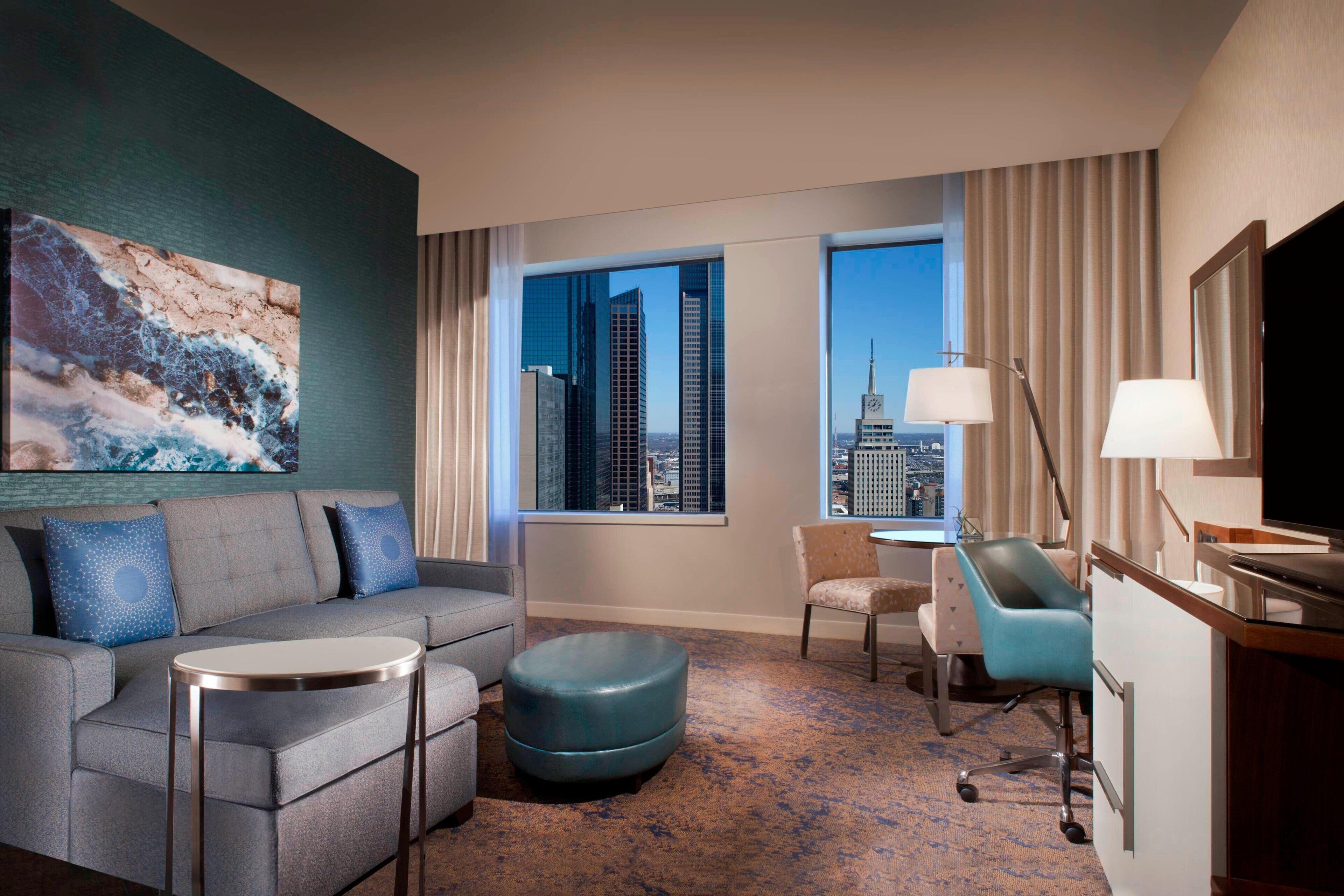 Habitación de hotel moderna con vista a la ciudad, un sofá, una mesita, una silla y un escritorio con una lámpara.