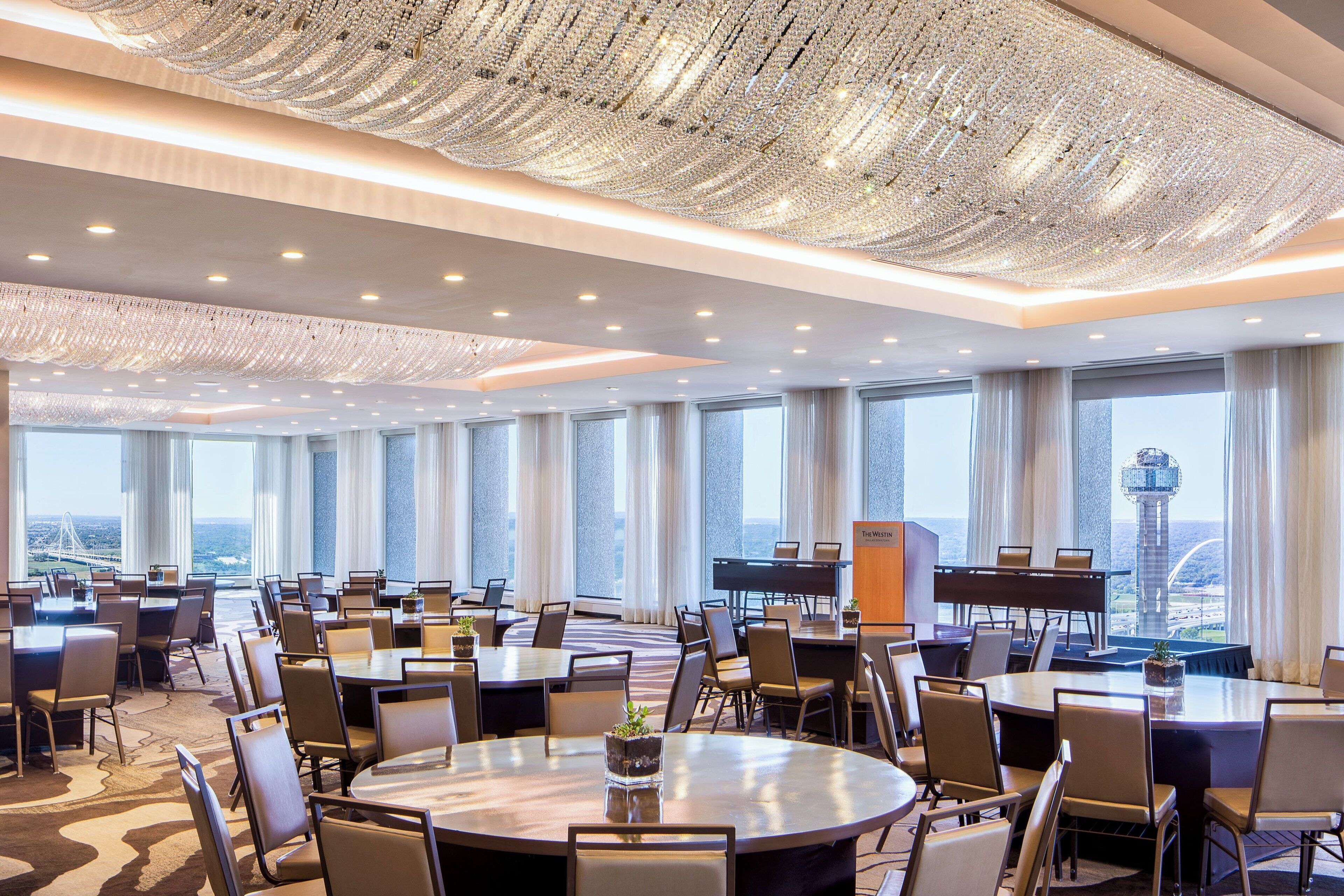 A spacious conference room with multiple tables and chairs, large windows with a view of a cityscape, and a decorative ceiling.