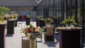 An outdoor seating area with tables and chairs, surrounded by large windows and potted plants. - The Westin Dallas Downtown (Dallas)