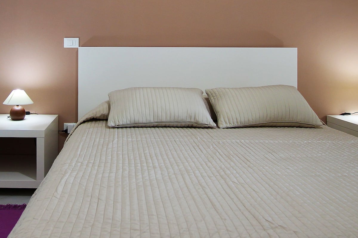 A neatly made bed with a beige comforter and pillows, flanked by two bedside tables with lamps, in a room with a simple, clean design.