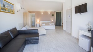 A modern hotel room with a sectional sofa, a bed with a headboard, a small desk with a TV, and a refrigerator. - Belvedere Rent Rooms (Campofelice di Roccella)