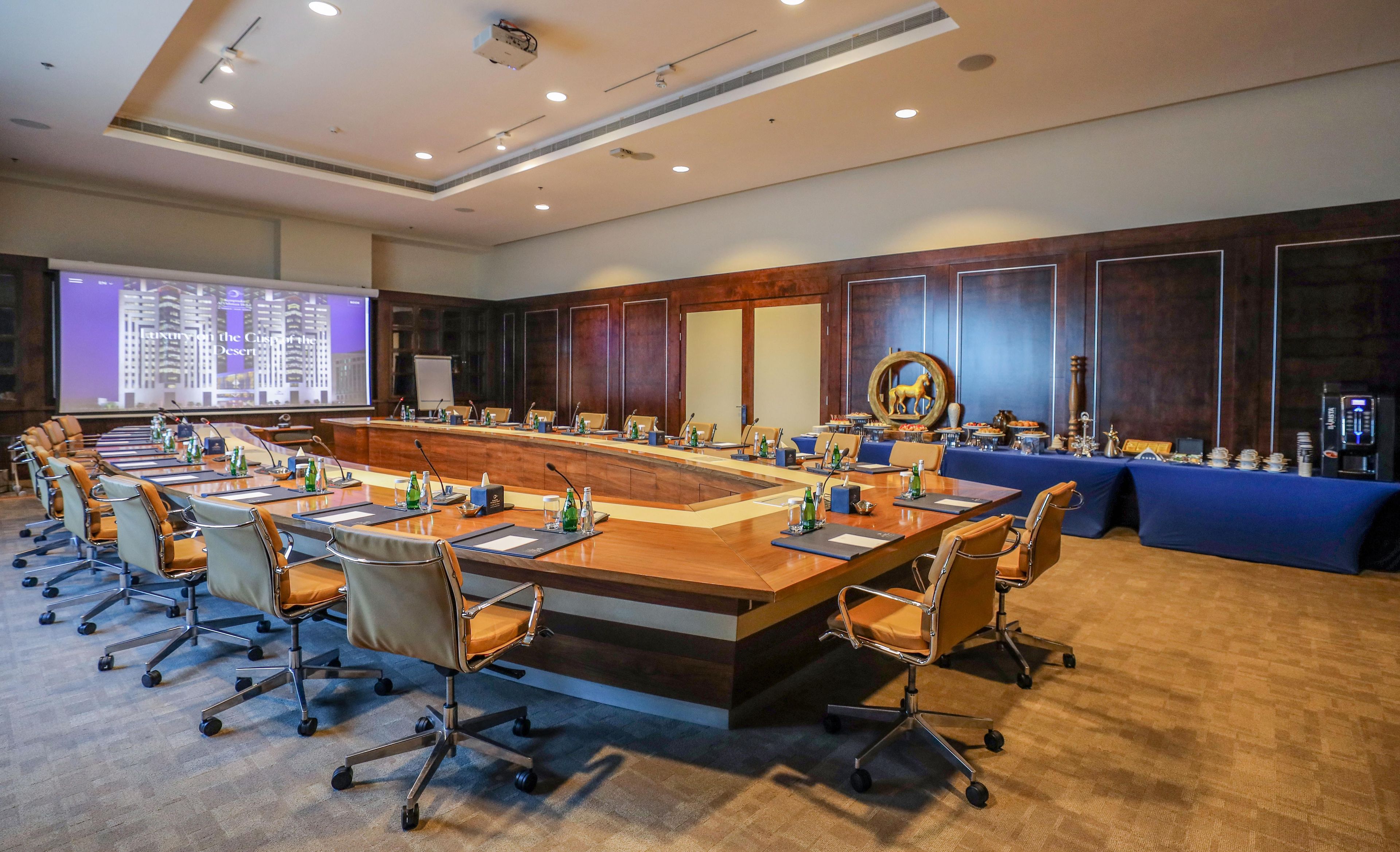 Un salón de conferencias con una larga mesa, sillas, una pantalla de proyección y un rincón con café.