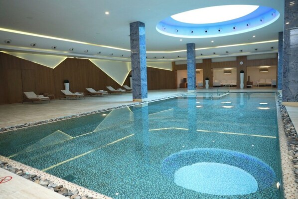 A modern indoor swimming pool with a geometric tile design, lounge chairs, and a circular skylight. - Kempinski Al Othman Hotel Al Khobar (Al Khobar)