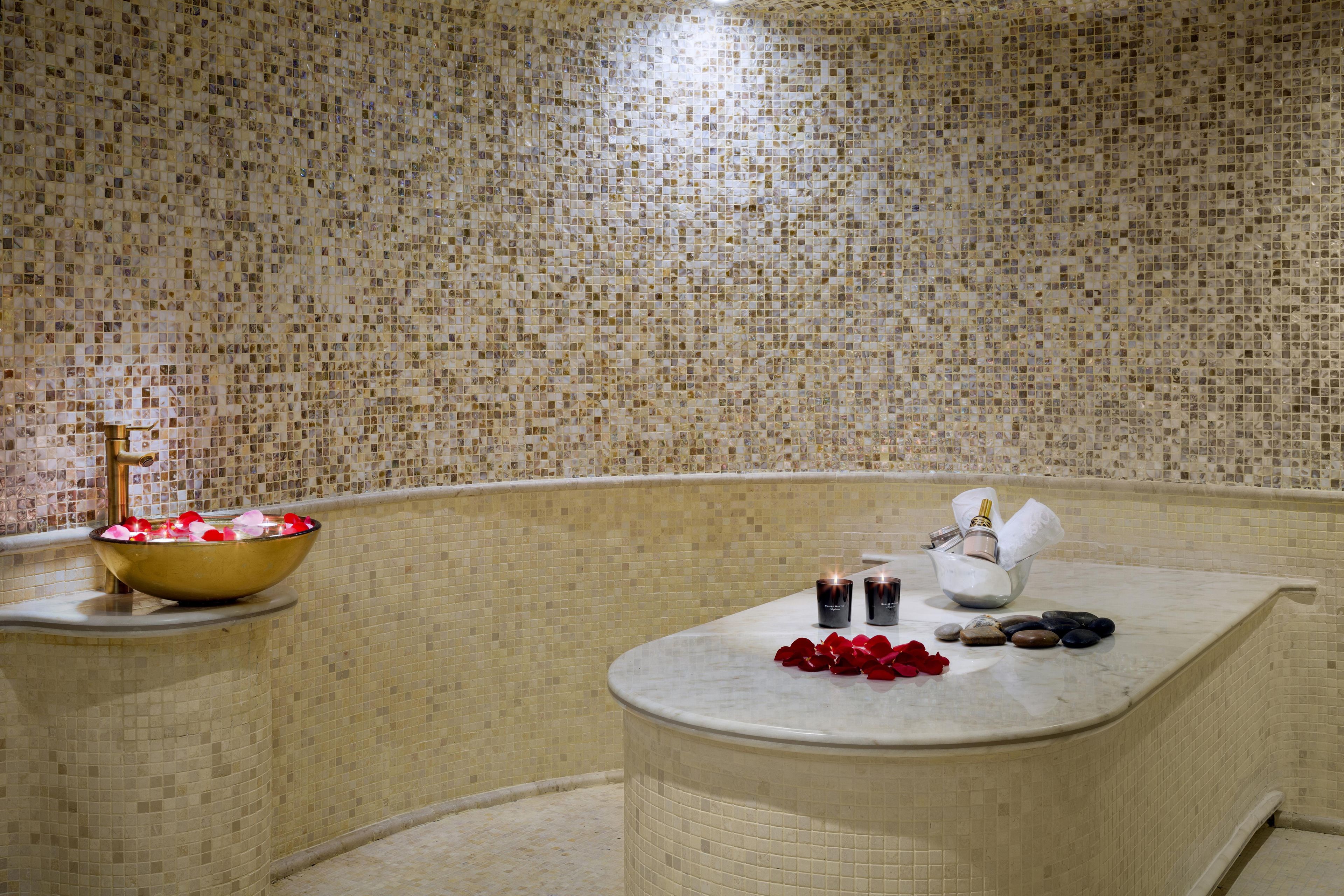 Un área de spa con una pared de mosaicos, un lavabo dorado con pétalos de rosa, velas y un recipiente de piedra.