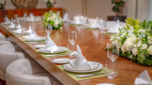 A long dining table set with white plates, folded napkins, and wine glasses, surrounded by chairs.
