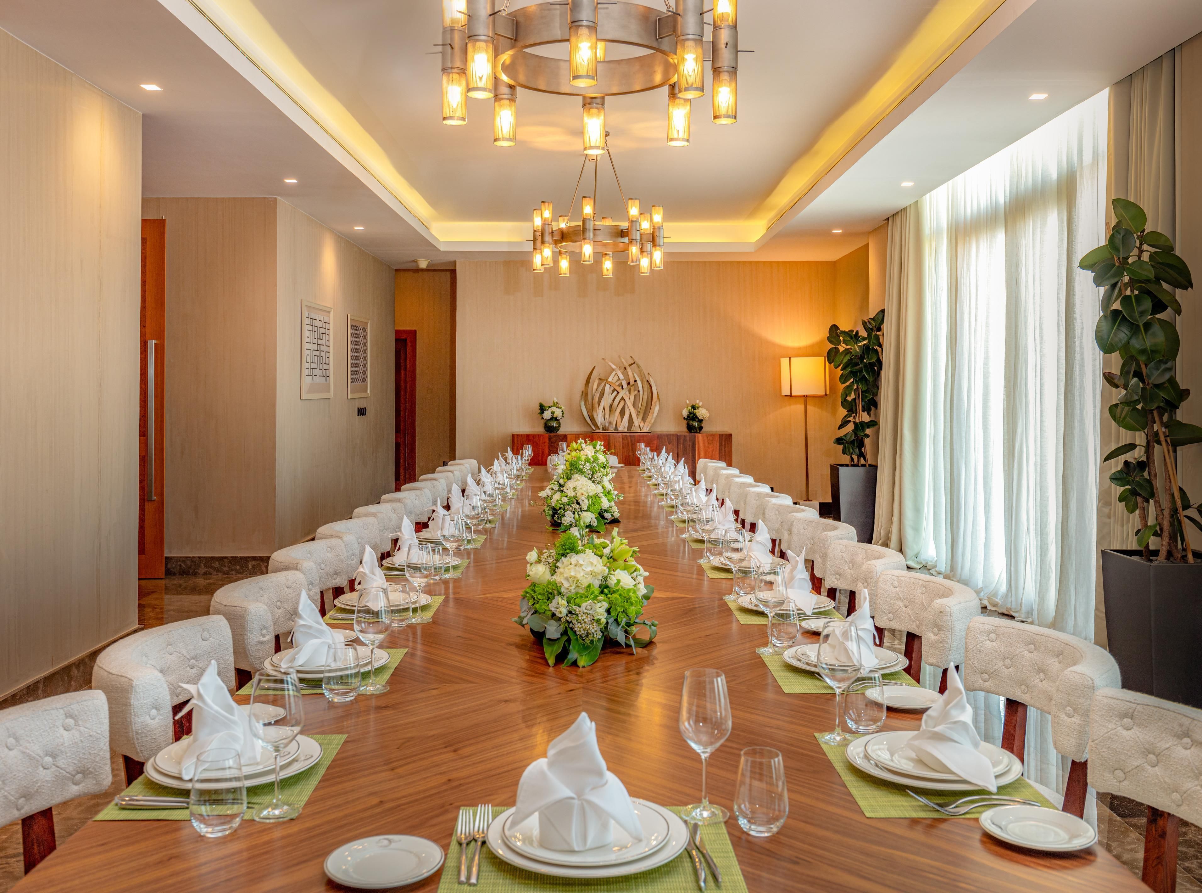 A conference room with a long table set for a formal event, surrounded by chairs, a chandelier, and a large plant.