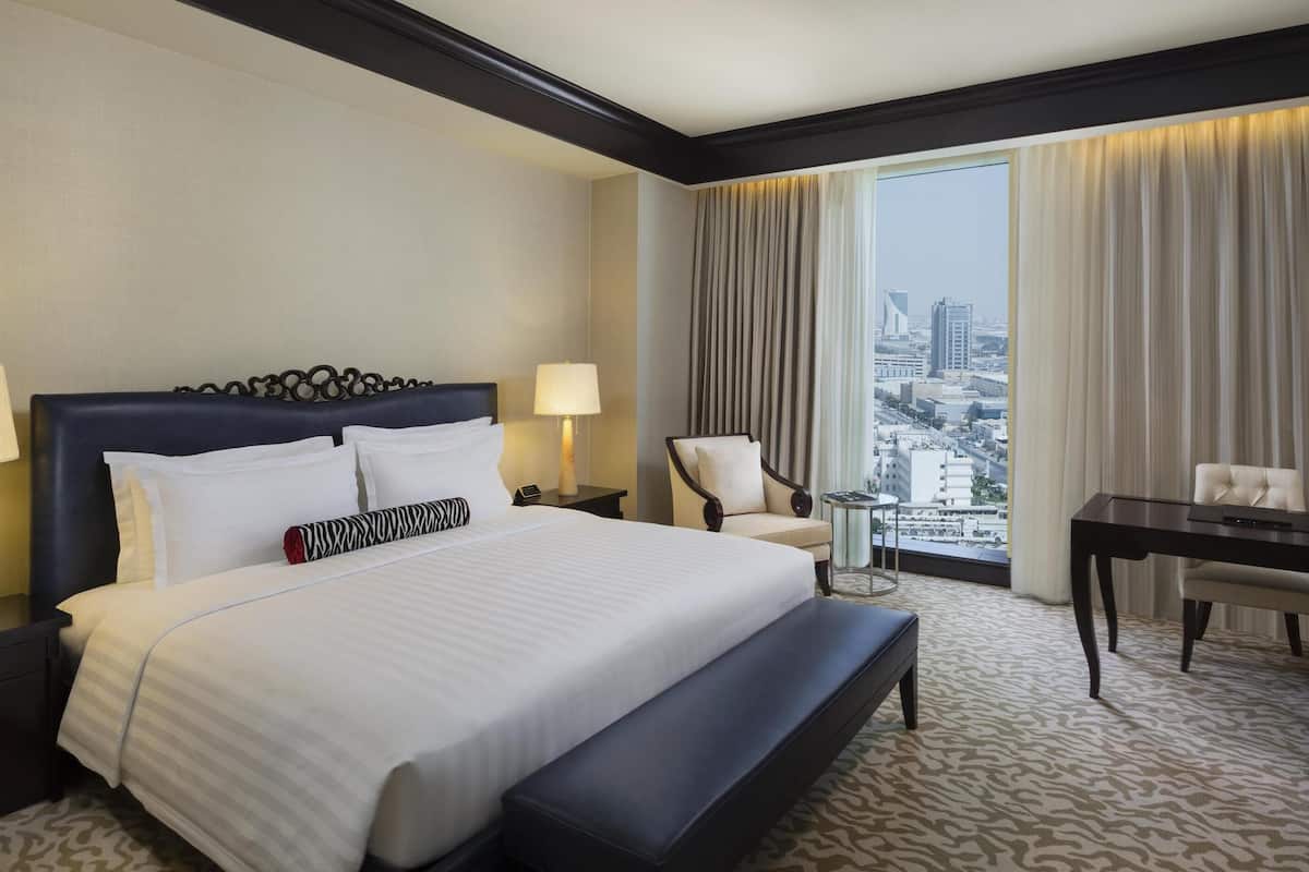 Ein Hotelzimmer mit einem großen Bett, Nachttischen, einem Schreibtisch, einem Sessel und Blick auf die Stadt.