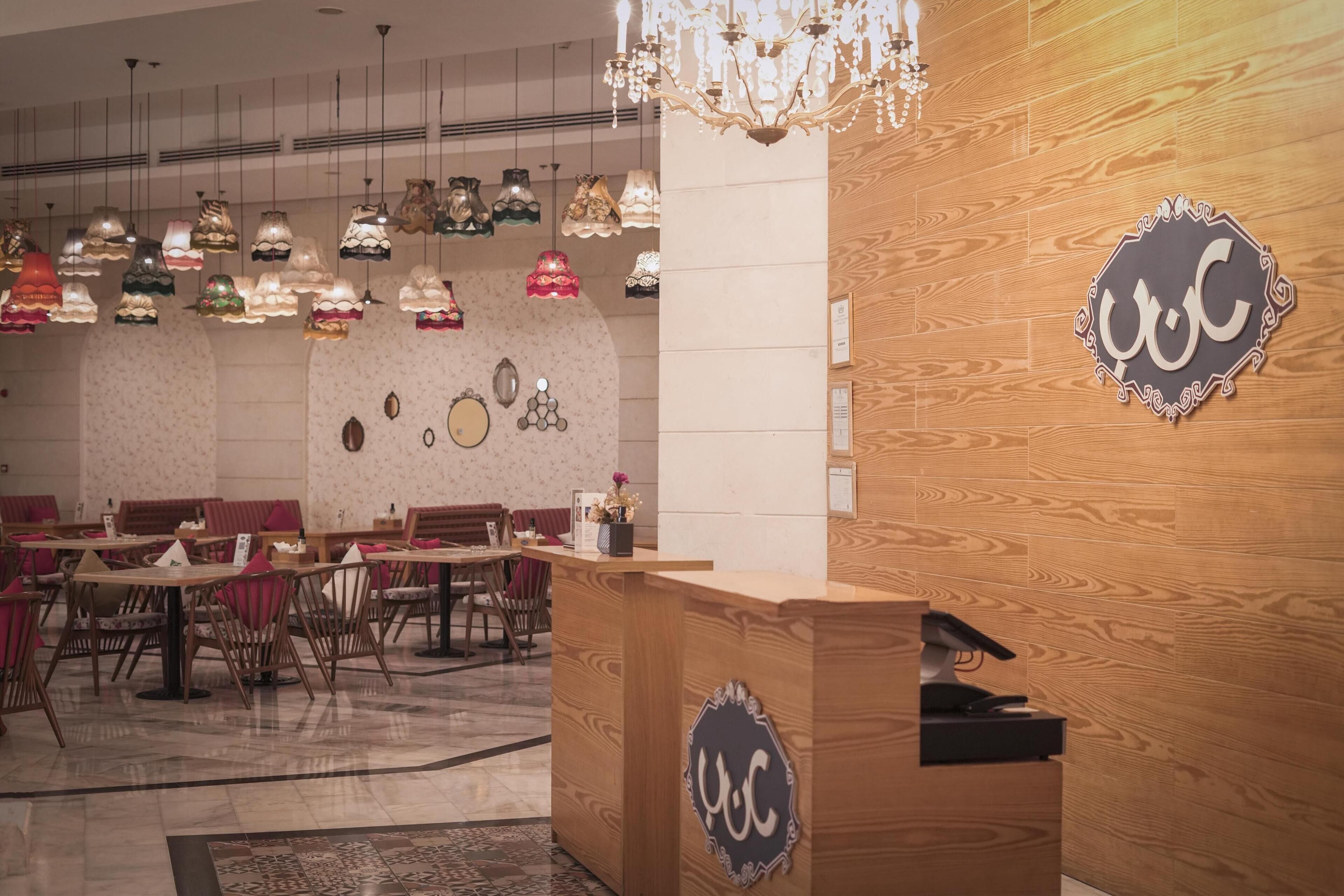 A restaurant interior with wooden reception desk, multiple dining tables, chairs, hanging pendant lights, and a chandelier.