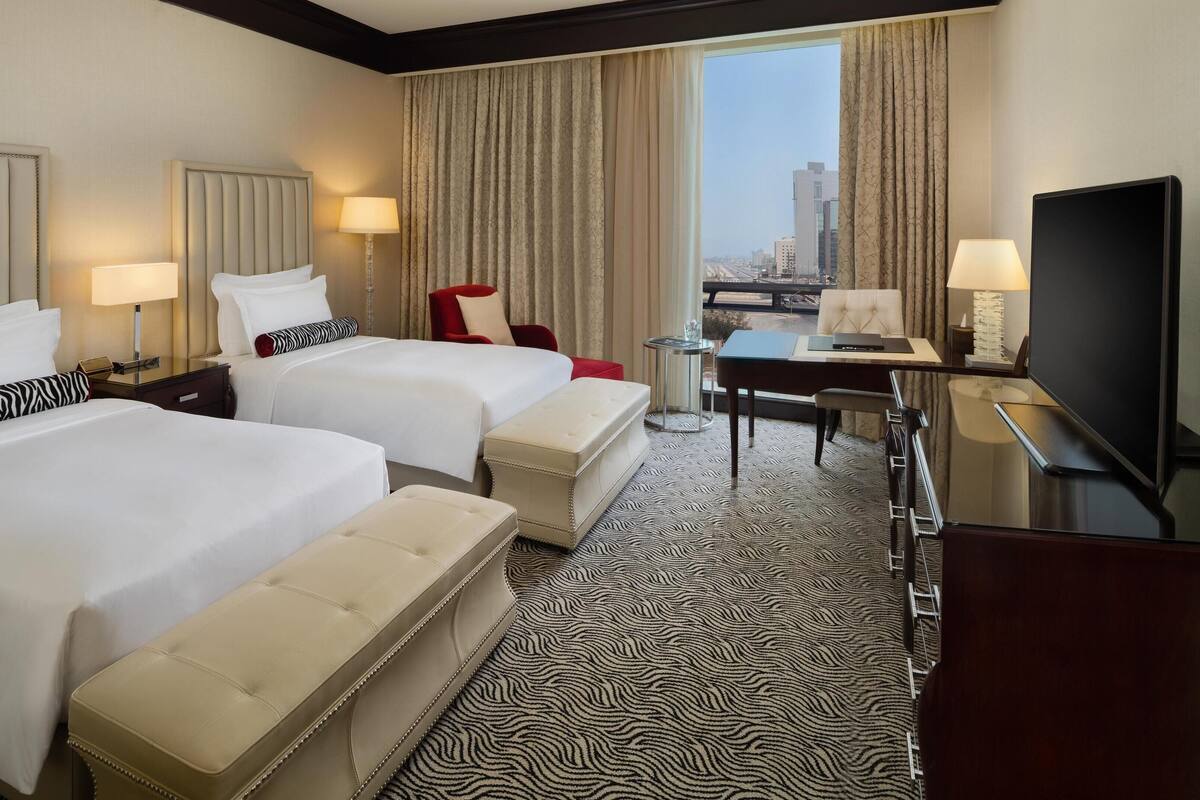 Ein Hotelzimmer mit zwei Betten, einem Schreibtisch, einem Fernseher und Blick auf die Stadt.