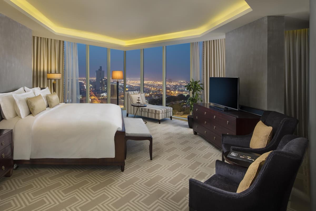 Ein Hotelzimmer mit einem großen Bett, einem Fernseher, einer Sitzecke und Blick auf eine nächtliche Skyline.