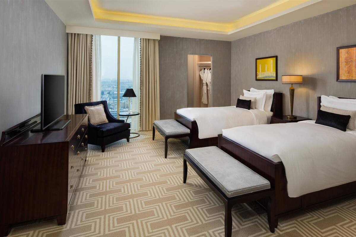 Ein Hotelzimmer mit zwei Betten, einem Schreibtisch, einem Sessel, einem Fernseher, einer Lampe und Blick auf die Stadt.