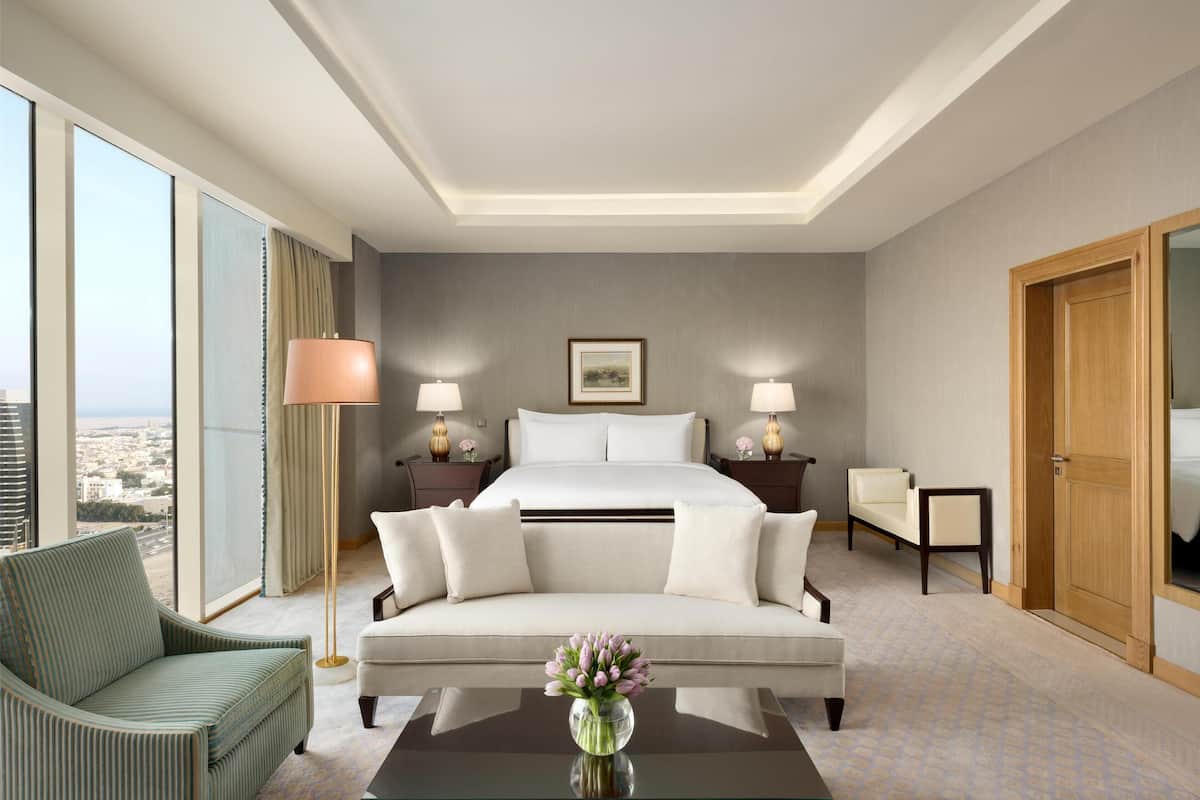 Ein Hotelzimmer mit einem großen Bett, einer Sitzecke mit einer Bank, einem Sessel, Nachttischen mit Lampen und Blick auf die Stadt durch große Fenster.