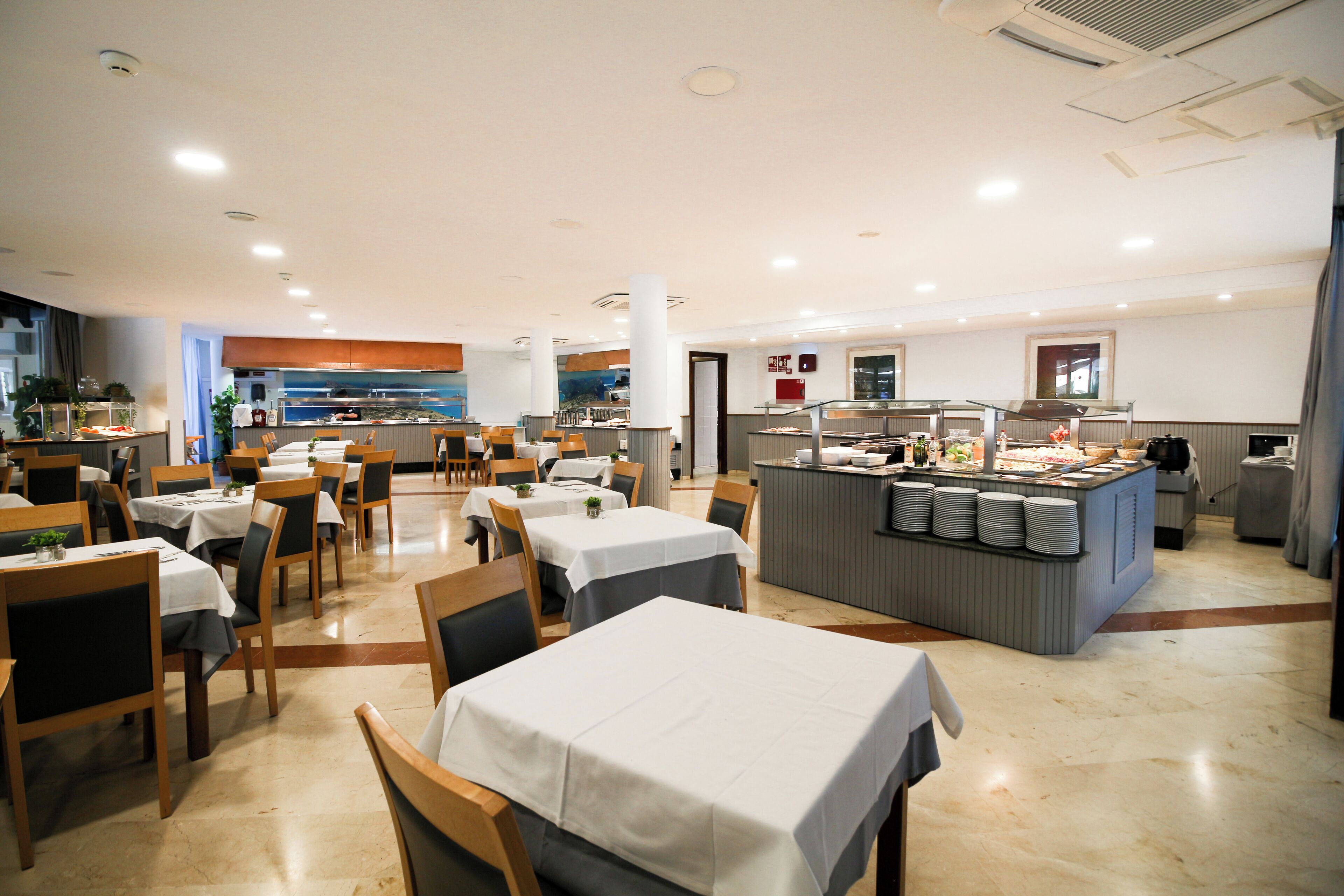 Un restaurant spacieux, doté de nombreuses tables et chaises, d’un espace buffet et d’un comptoir où sont présentés les plats.