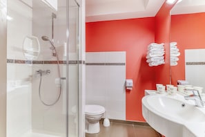 A bathroom with a glass-enclosed shower, a white toilet, and a sink with toiletries. - ACE Hôtel Montluçon (Saint-Victor)