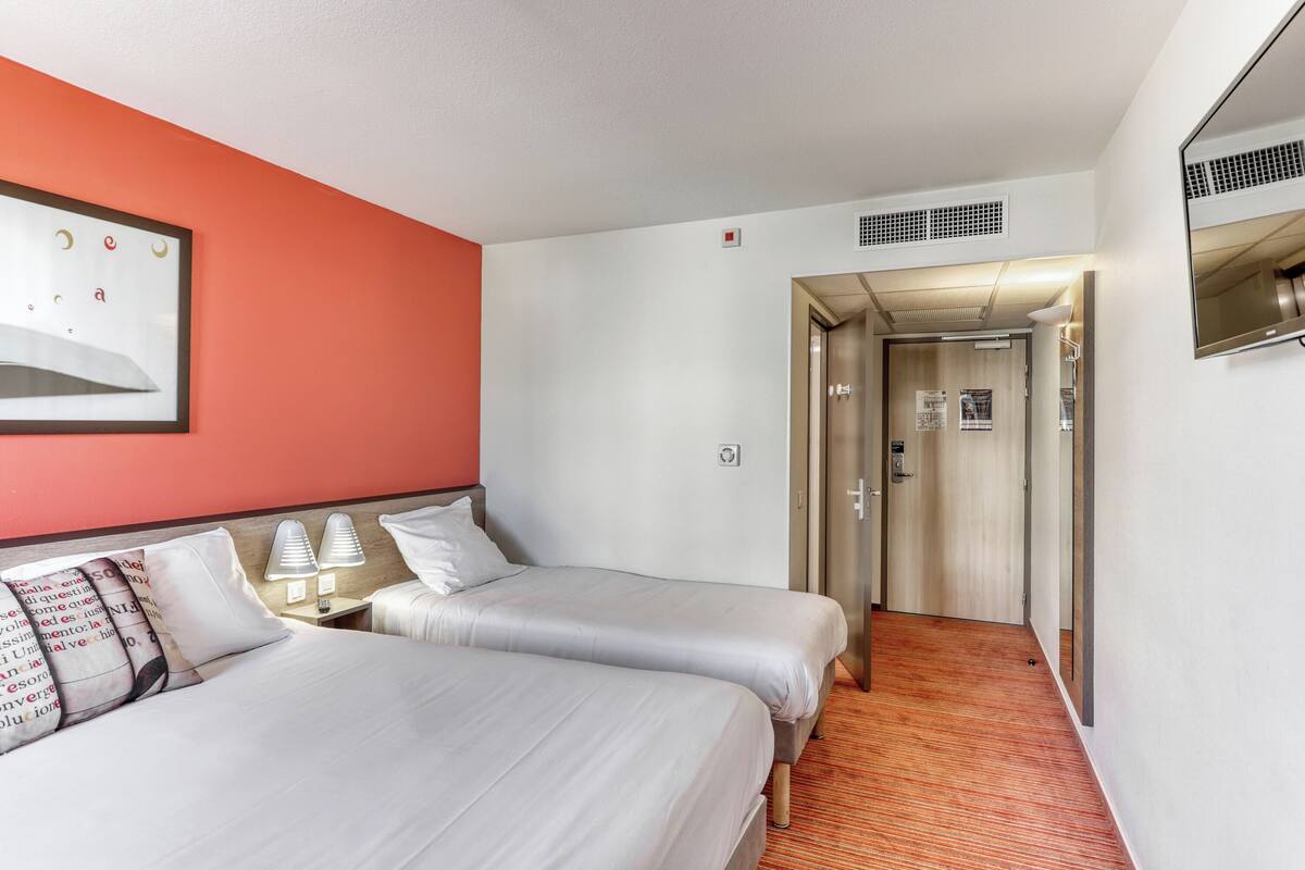 A hotel room with two beds, a red accent wall, a framed picture, a mirror, and a door leading to a hallway.