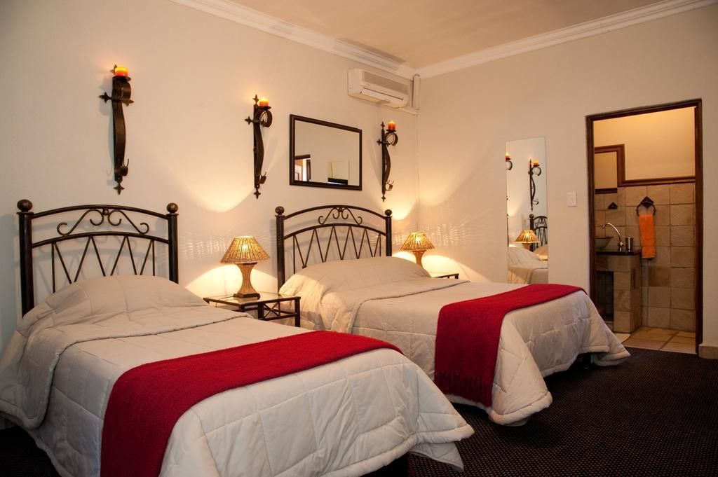Habitación de hotel con dos camas, cada una con una manta roja, mesitas de noche con lámparas y apliques de pared.