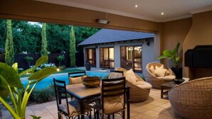 A poolside area with wicker furniture, a dining table, and a small building with a tiled roof. - Rise and Shine Lodge (Bloemfontein)