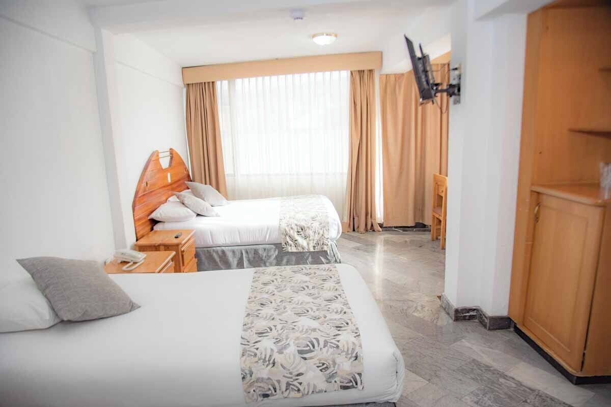Habitación de hotel con dos camas, un armario de madera, un escritorio con teléfono y un ventanal con cortinas.