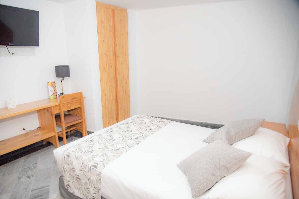 Un dormitorio con cama, escritorio, silla, televisor montado en la pared y panel de madera en la pared.